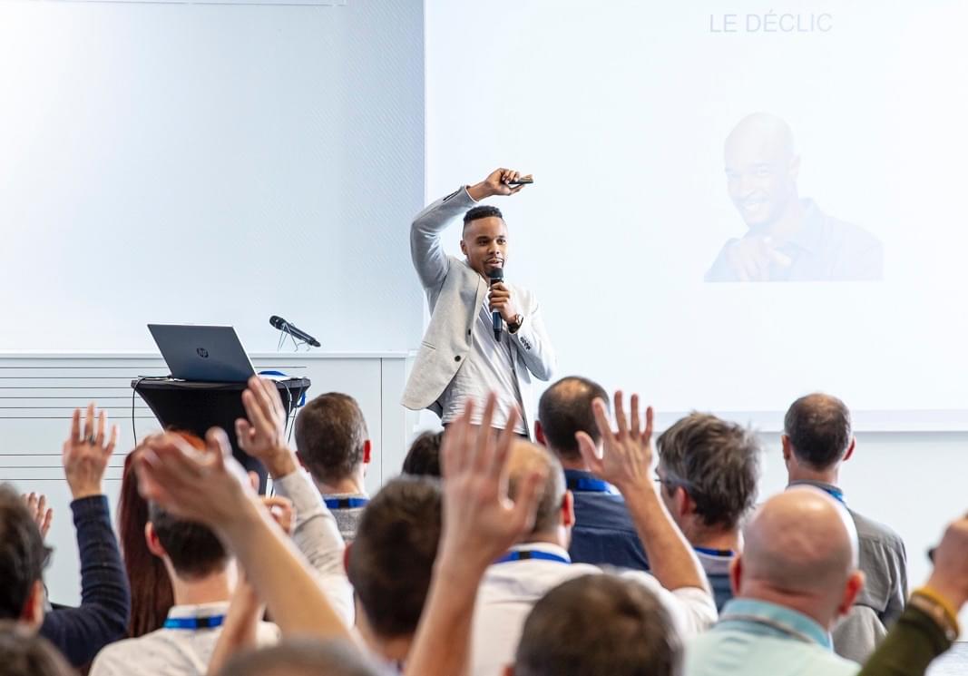 Steven sur scène en conférence, micro à la main, face à une salle aux mains levées